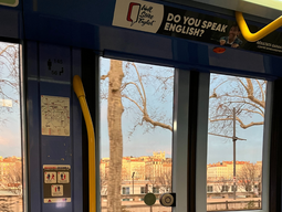 Inside a tram, the view through large windows reveals a tree-lined street with ornate buildings bathed in sunset light. Yellow handrails and an English ad are visible.