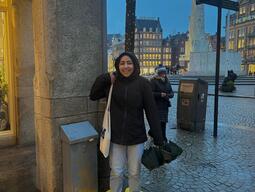 A person smiling in oversized yellow clogs on a wet city street at dusk, adjacent to a large stone monument. The scene conveys humor and whimsy.