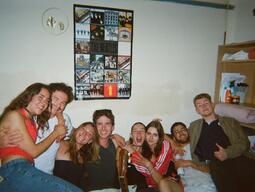 A group of eight friends smile and pose energetically on a couch in a cozy room. A Beatles poster hangs above them, adding a cheerful, nostalgic vibe.