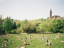 Sunny park scene with people sunbathing on a grass lawn, surrounded by lush green trees. A historic building with a tall tower is visible in the background.