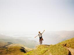 A person stands joyfully on a grassy mountain peak, arms outstretched, overlooking a vast, serene landscape with lakes and distant hills under a clear sky.
