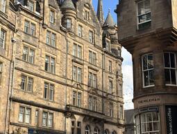 Historic street scene with ornate sandstone buildings under a blue sky. People walk below, adding liveliness to the charming, architectural setting.