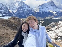A cheerful duo takes a selfie with snow-capped mountains in the background. Bright sky and dramatic peaks complement their joyful expressions.