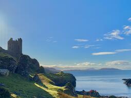 A medieval stone castle towers over a grassy cliff, overlooking a calm blue sea under a bright, clear sky. The scene is serene and picturesque.