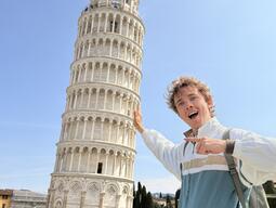 A person playfully poses as if holding up the Leaning Tower of Pisa. They smile excitedly on a sunny day, with tourists milling around in the background.