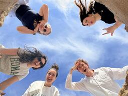 Five people smiling and posing while looking down into a sand pit, with a blue sky above. The scene conveys a playful, joyful atmosphere.