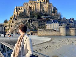 Person in a coat gazes at Mont Saint-Michel, France, under a clear blue sky. The historic abbey stands majestically on a rocky hilltop.