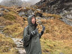 A person in a rain poncho smiles and gestures with excitement while hiking on a misty, rocky trail. The terrain is covered in wet, orangey-brown grass.