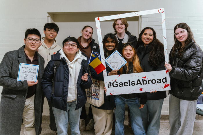 A diverse group of eight people smiling, holding a #GaelsAbroad frame. They wear casual winter clothing, exuding a joyful and inclusive atmosphere.