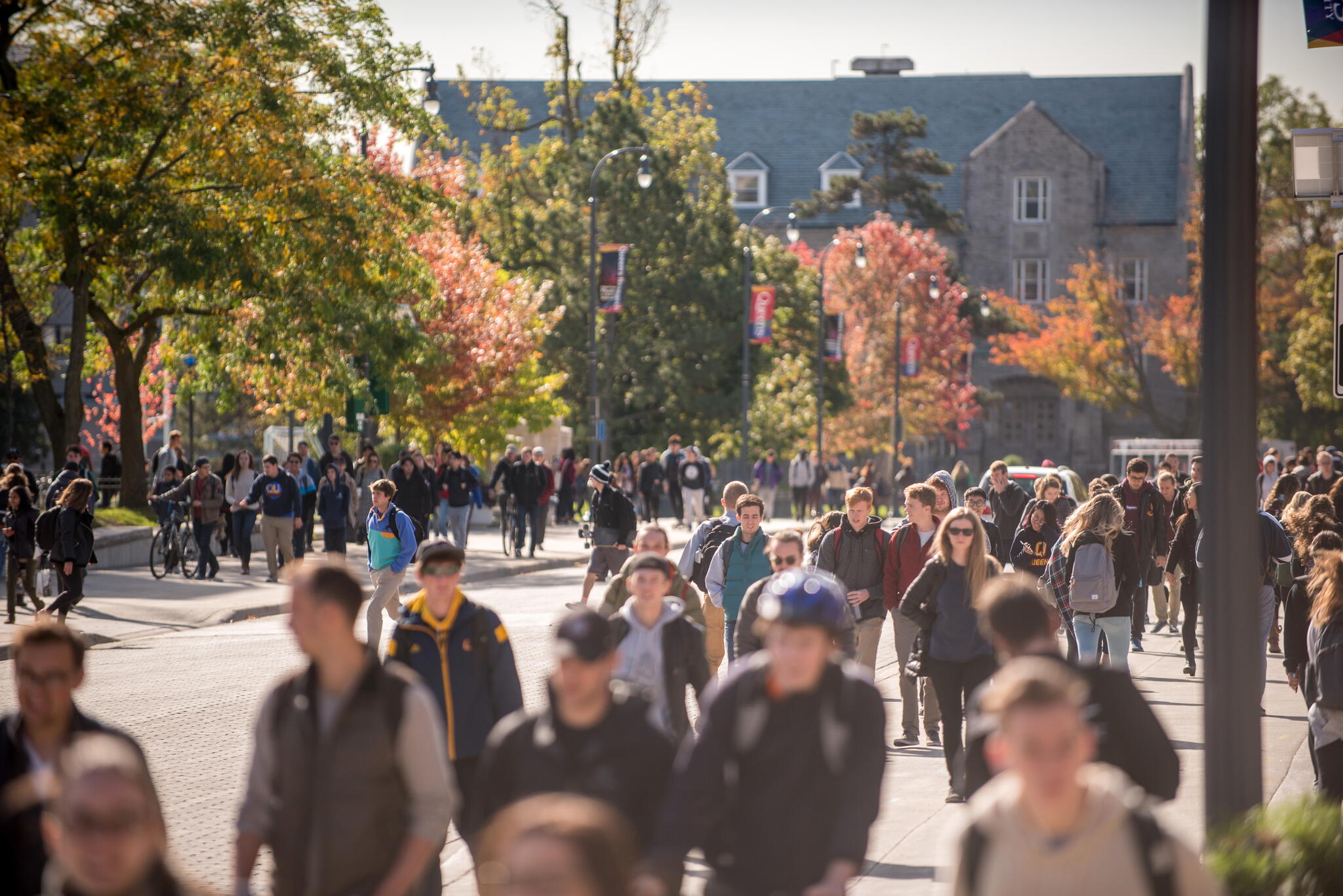 Students walk along a busy university campus pathway lined with trees showing autumn colours, with academic buildings visible in the background.