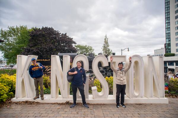 three students standing in front of the Kingston sign located downtown Kingston. 