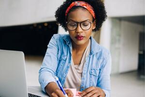a woman sits at a desk in front of a laptop and writing on a notepad