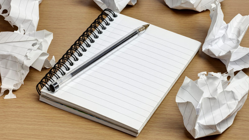 A blank spiral notebook with a pen resting on it, surrounded by crumpled pieces of paper on a wooden desk.