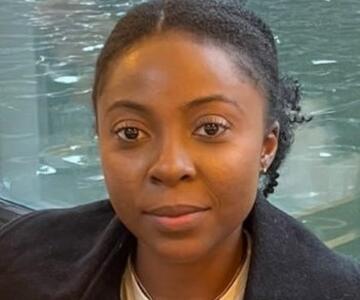 Patricia Ackah-Baidoo, wearing a black jacket, white shirt, sitting in front of a body of water