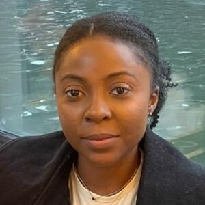 Patricia Ackah-Baidoo, wearing a black jacket, white shirt, sitting in front of a body of water
