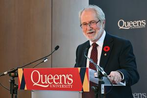 Patrick Deane speaks into mircophone at podium with Queen's logo 