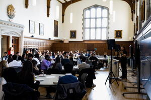 Large group of students wearing professional clothing sitting in building facing a woman giving a speech in front of a screen with a slideshow playing.