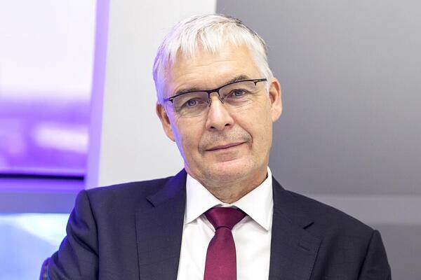 Man with white hair and glasses in a suit looking at camera.
