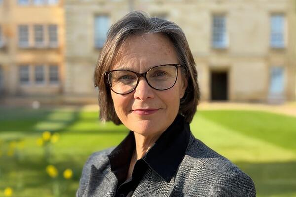 Woman with short hair wearing glasses and a black suit looking at camera with blurred limestone building background.