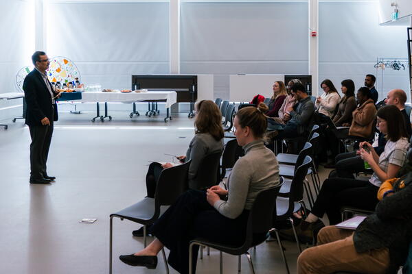 Man in black suit speaking and standing in front of an audience that is sitting in chairs, taken from the side.