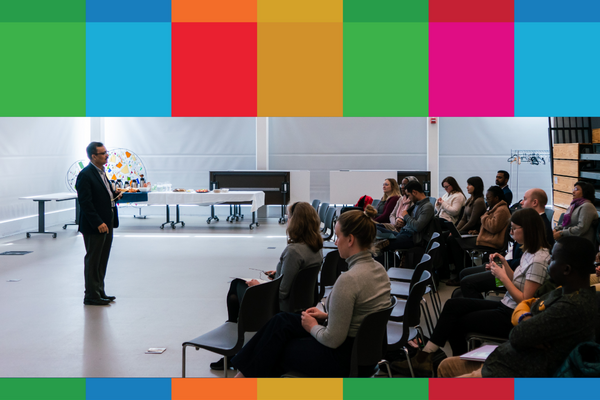 Man in black suit speaking and standing in front of an audience that is sitting in chairs, taken from the side.