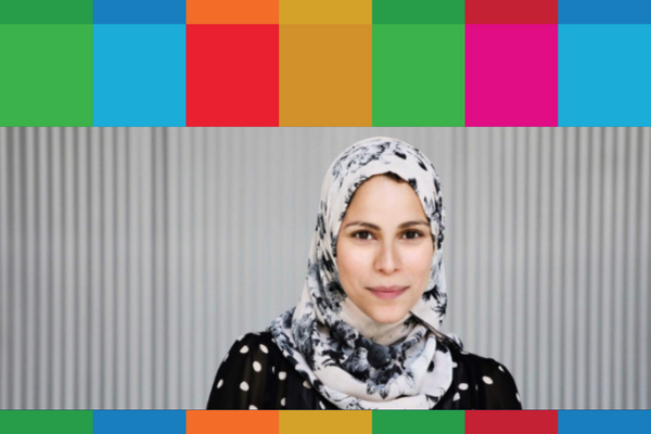 Woman with white and grey watercolour hijab and a black shirt with white polka dots standing in front of a grey wall.