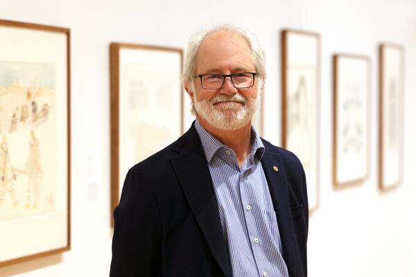 Man in blue shirt and dark blue blazer with glasses and white hair standing in front of framed artwork