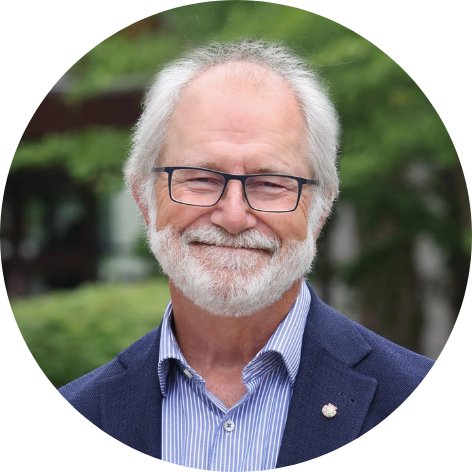 Man with white head wearing a blue suit, standing in front of blurred nature background, smiling at camera.