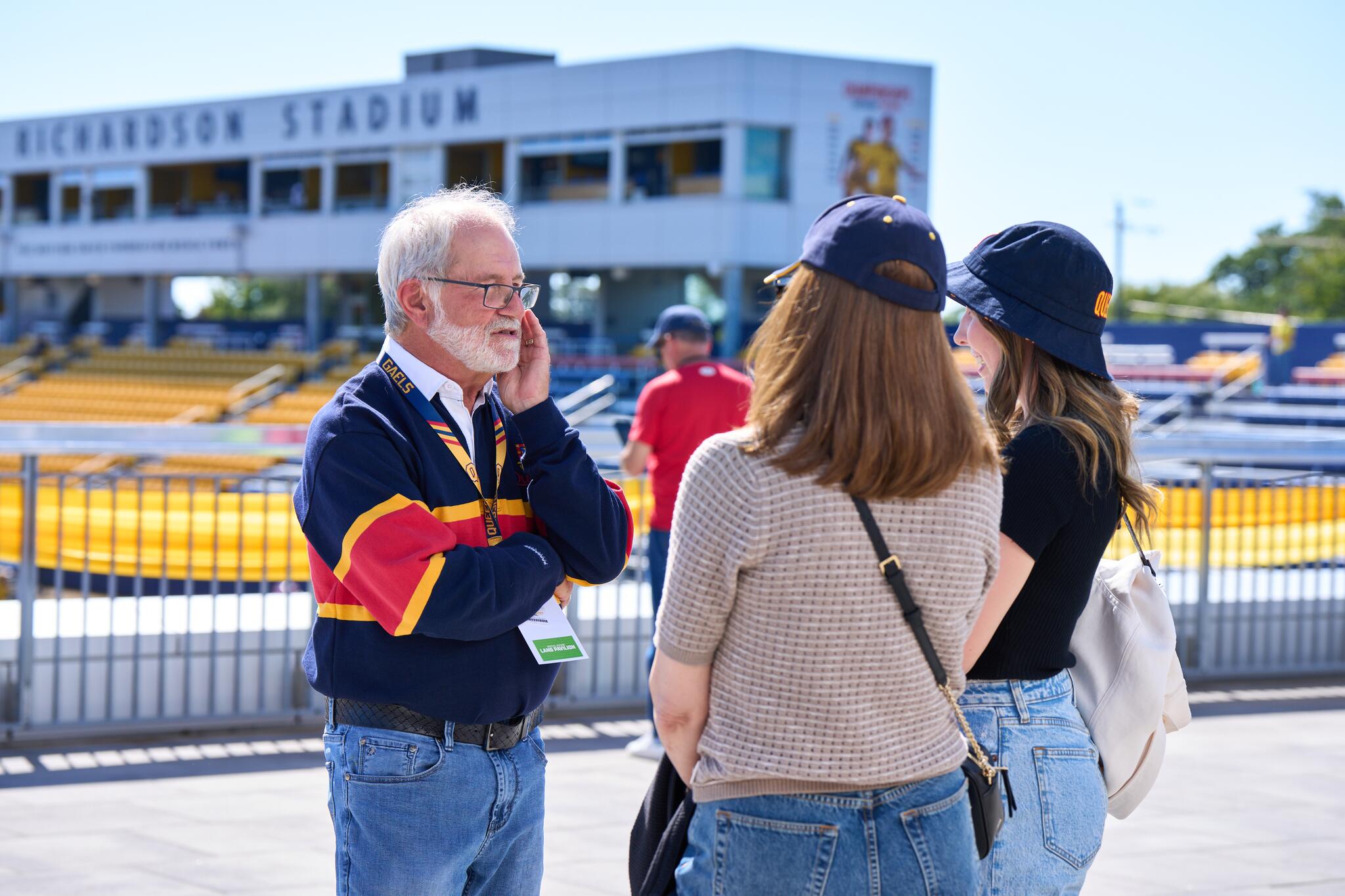 Patrick Deane, man with white hair wearing a rugby shirt in a blue, red and gold colourway, speaking to two women whose faces are not shown.