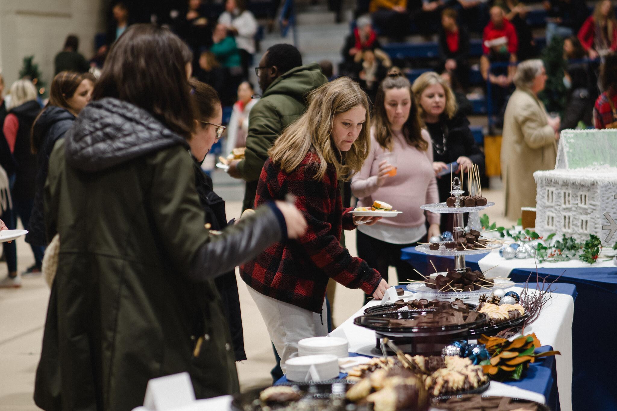 Women grabbing deserts from table covered by blue table cloth, in a gymnasium.