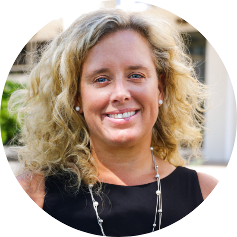 Woman with blonde curly hair, wearing a black dress and silver necklace, smiling at camera.