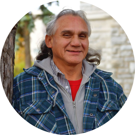 Man with long white hair, wearing blue gingham print jacket, leaning against a tree smiling at camera.