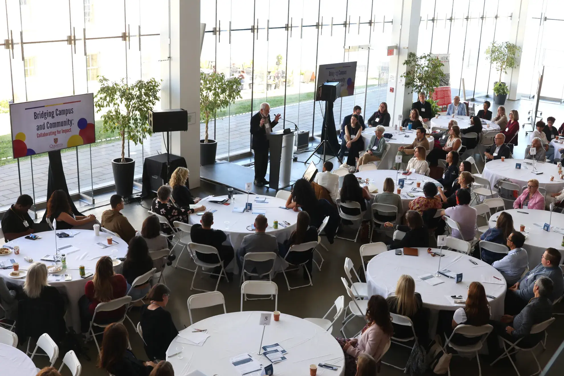 Aerial view of people sitting at circle tables in room with glass walls looking out to a lake.