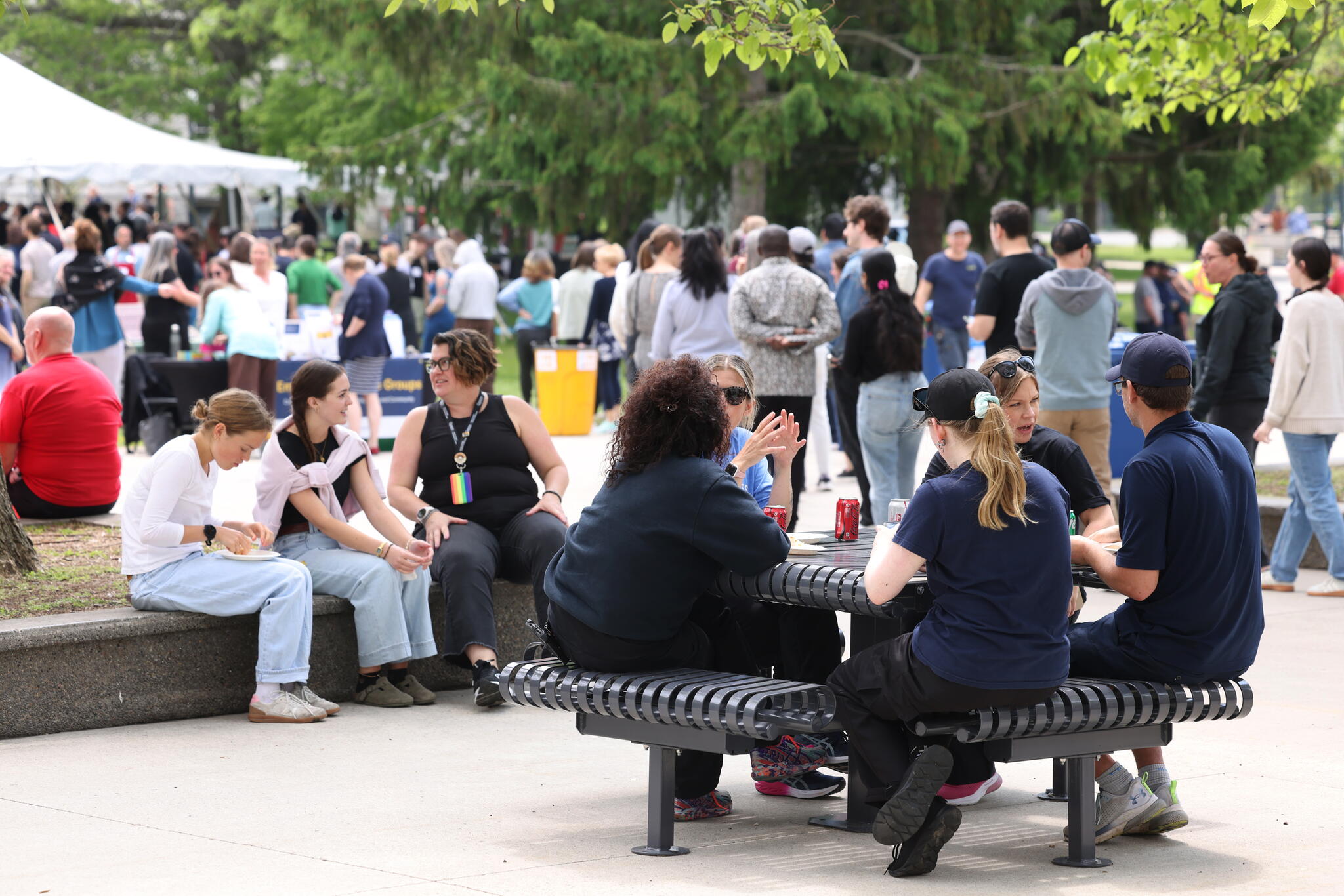 People sitting at a picnic table eating food and chatting, with large event tents and a lot of people in the background.