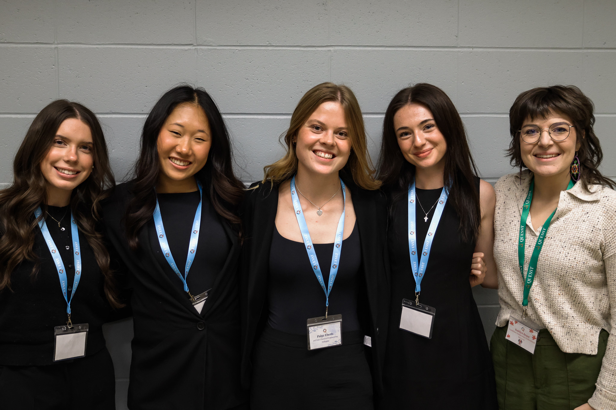 A group of five women smiling in front of a grey brick wall. The four on the left are of university age and are wearing all-black professional outfits. The woman on the right end is middle aged and is wearing a light-coloured business casual outfit.