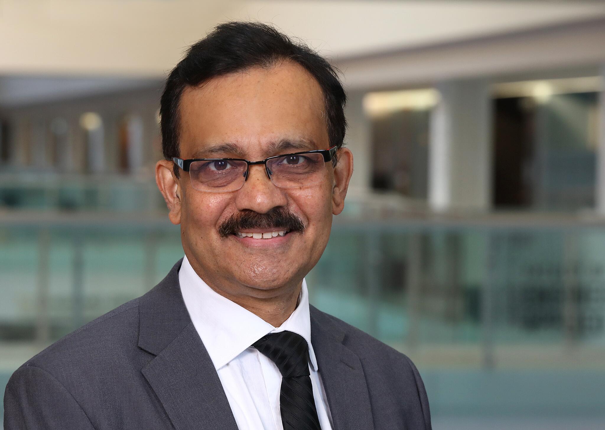 Man with brown skin with black moustache and hair wearing a suit and glasses.