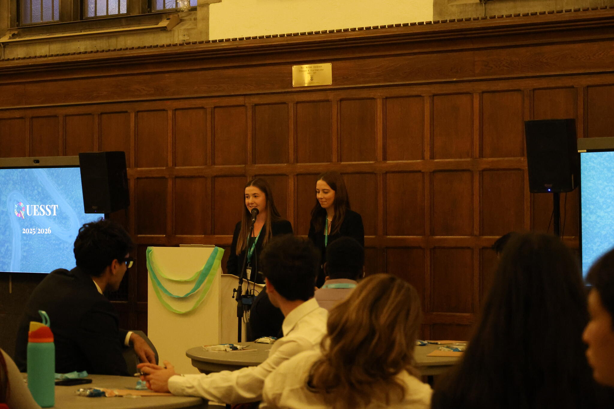 Two women, wearing professional clothing and lanyards, standing in front of a podium in a warmly lit room.