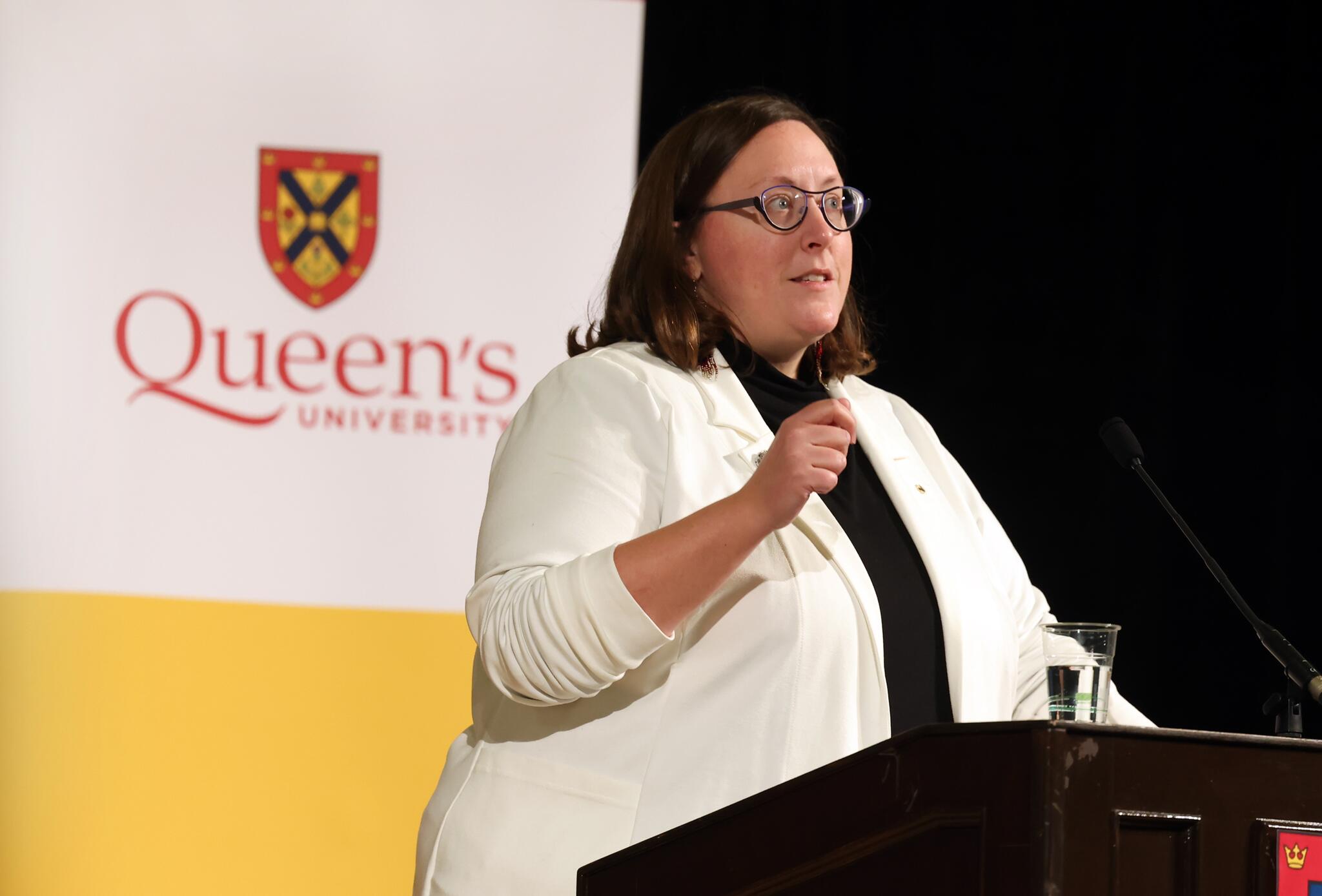 Woman in a white coat standing in front of wooden podium with the Queen's University Crest on the front.