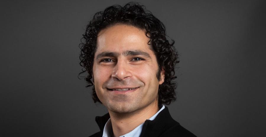 Man with black curly hair wearing a black quarter zip over a blue button up, smiling.