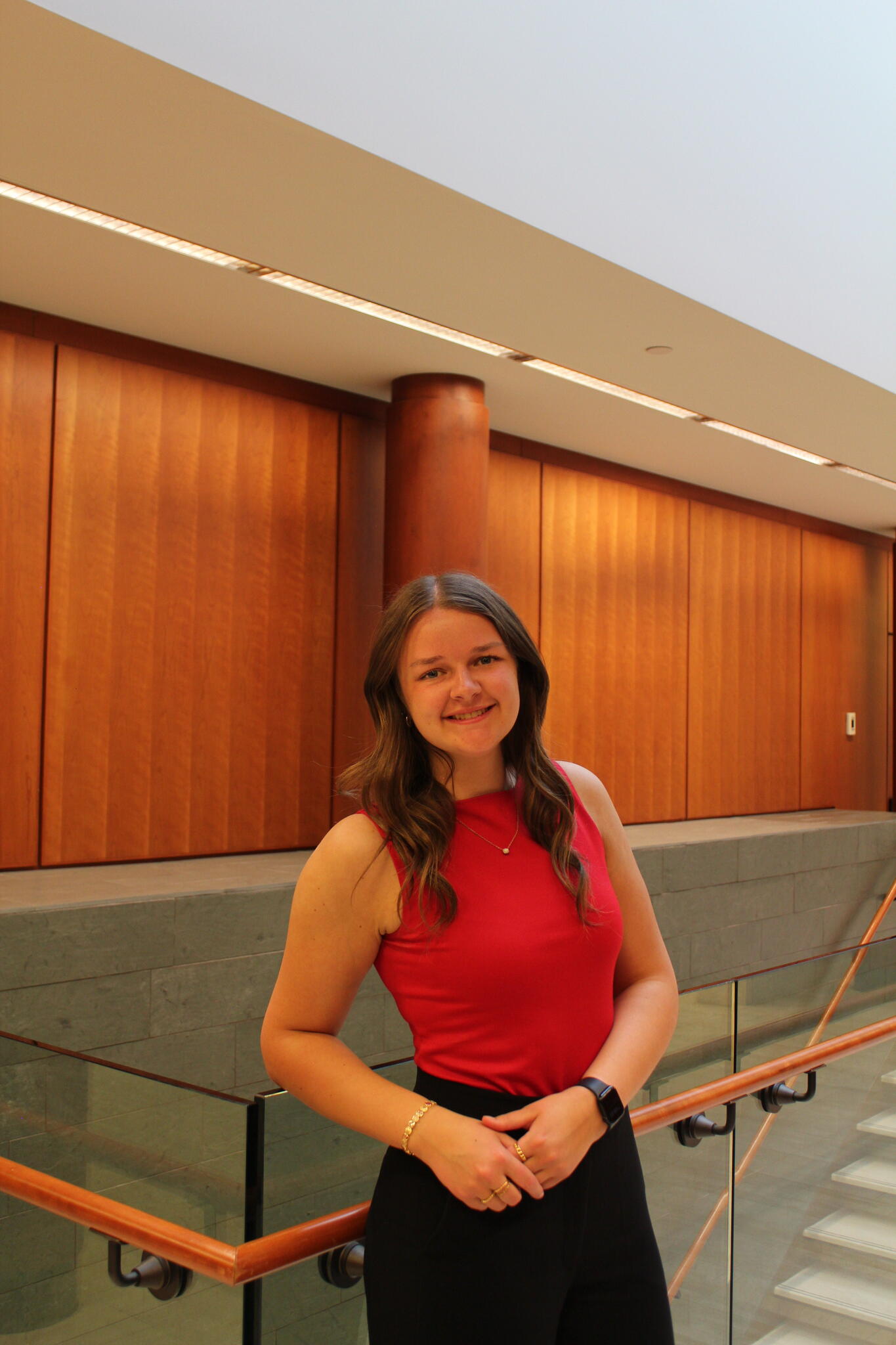Woman with long wavy brown hair wearing red blouse and black dress pants leaning against glass ceiling in modern wood-panelled building.