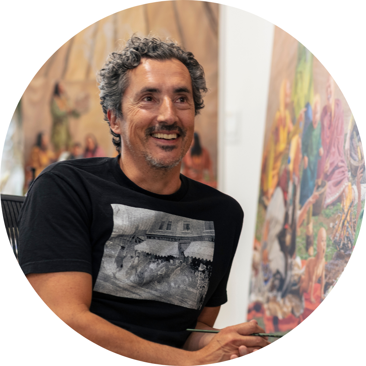 Man with curly black and grey-speckled hair wearing a black short sleave button up, standing in front of a colourful painting.