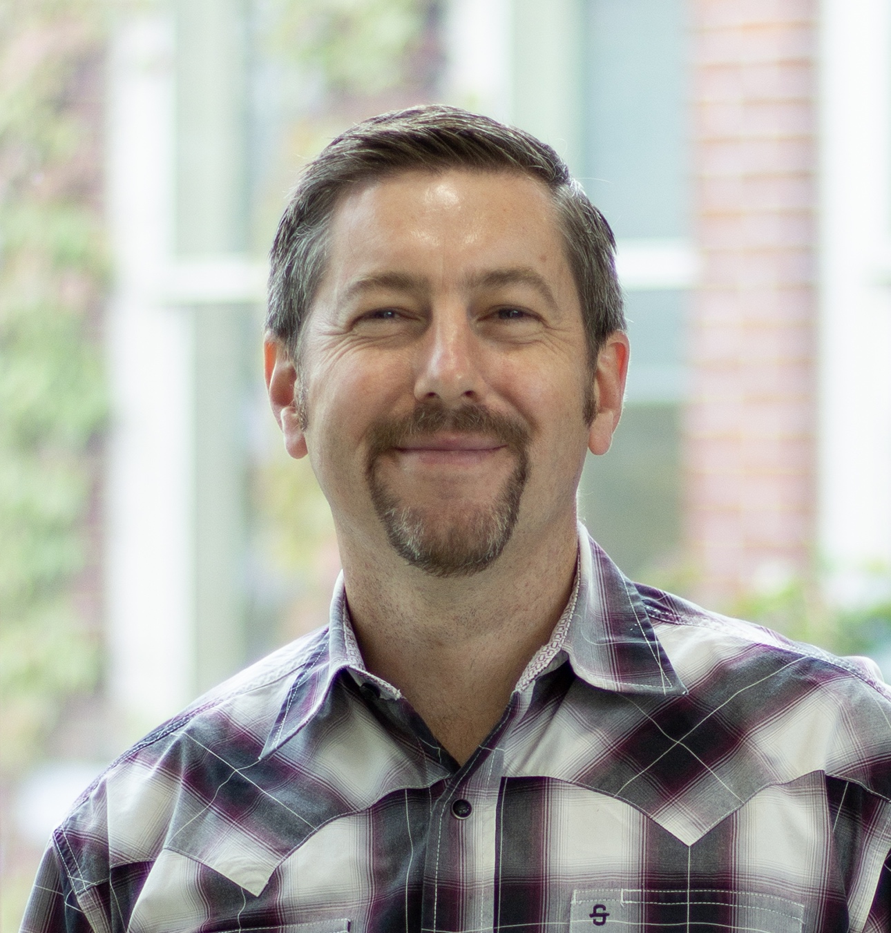 Man with grey hair and trimmed beard, wearing a red, blue and white plaid shirt, smiling at the camera.