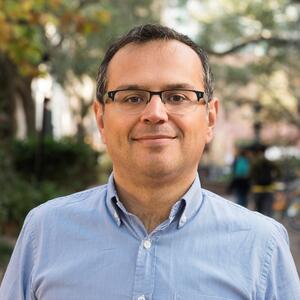 Man with brown skin and grey and black hair, wearing glasses and a blue button up, smiling.