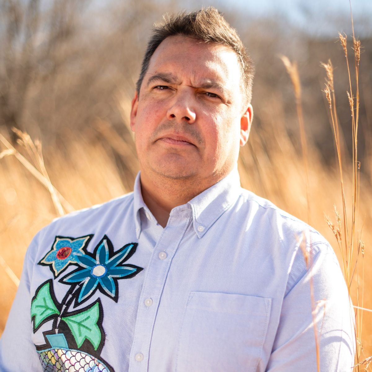 Man with light brown hair, wearing a light blue button up with indigenous beadwork on the left side, in front of a wheat field.