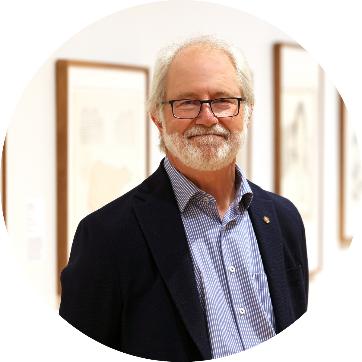 Man with white hair and beard, wearing a navy suit with a light blue button up, standing in front of a gallery of art.