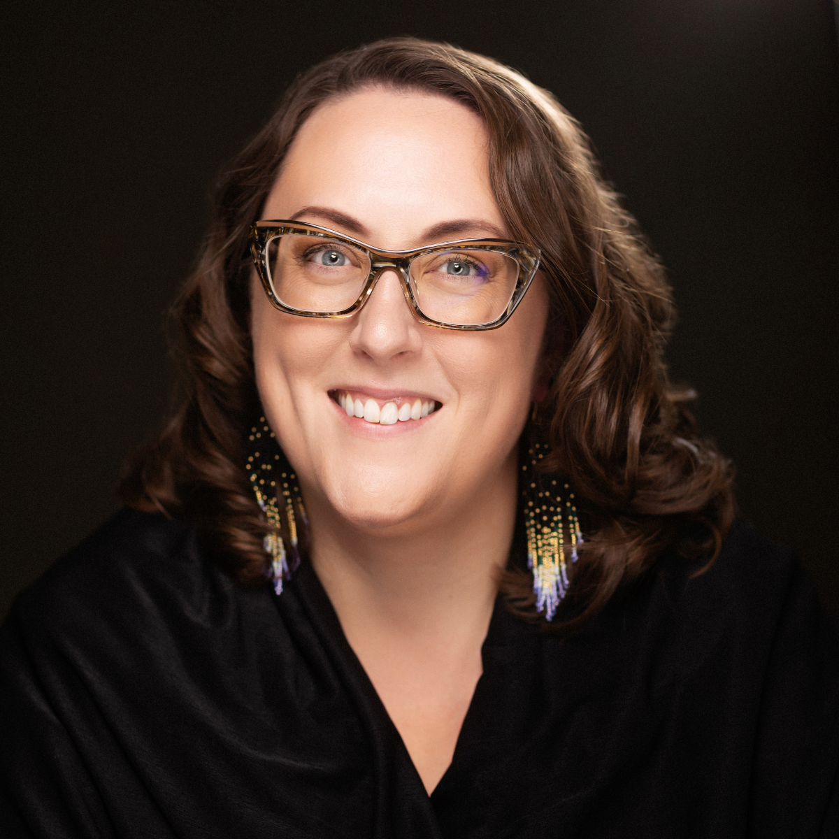 oman with brown hair, wearing a black shirt and big square glasses, smiling in front of a black backdrop.