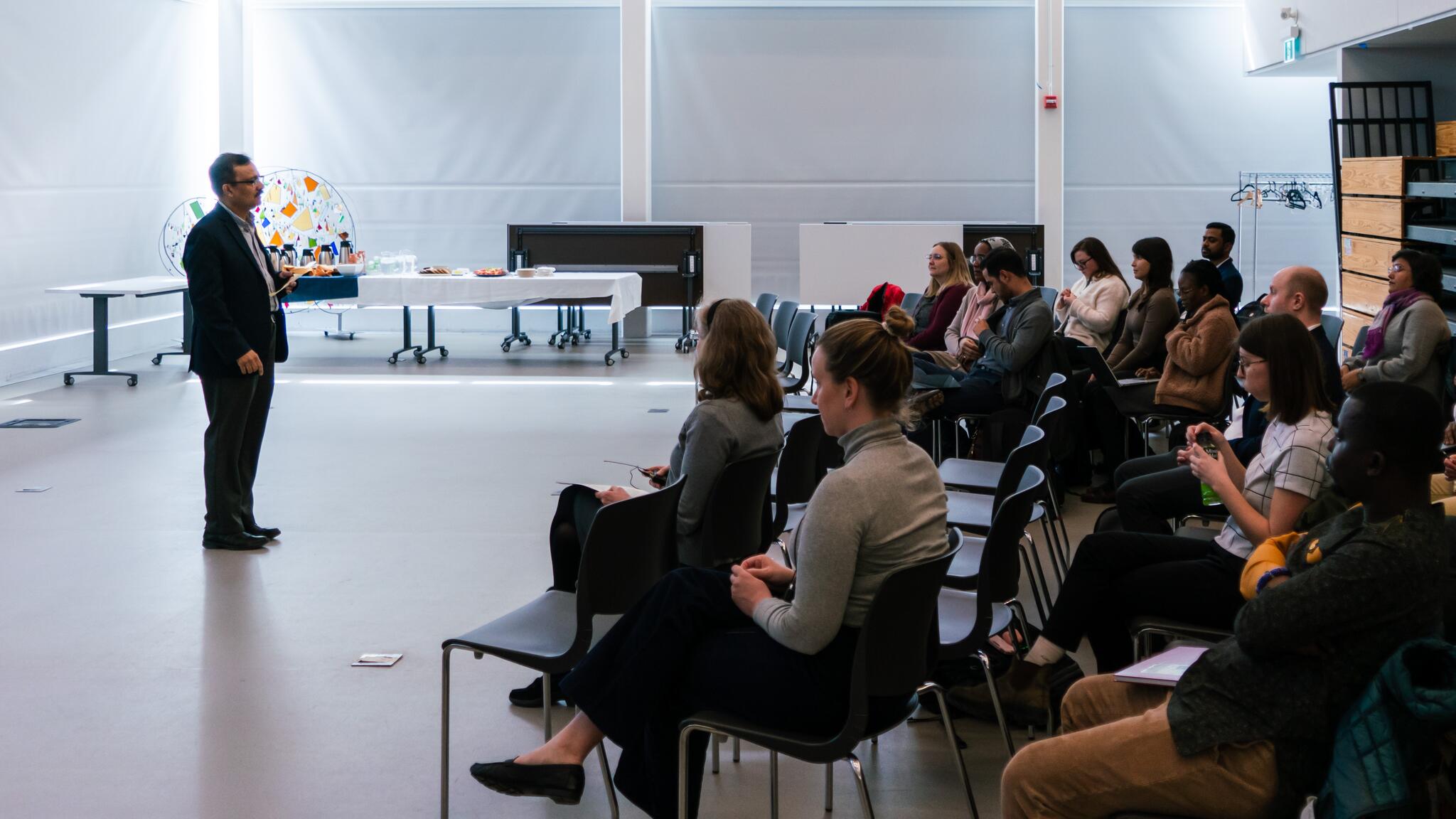 Man in black suit speaking and standing in front of an audience that is sitting in chairs, taken from the side.