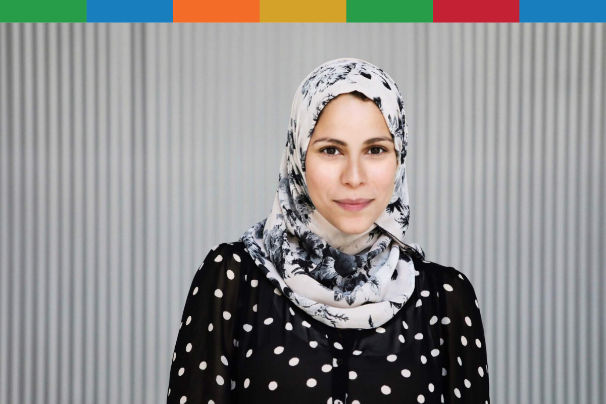Woman with white and grey watercolour hijab and a black shirt with white polka dots standing in front of a grey wall.