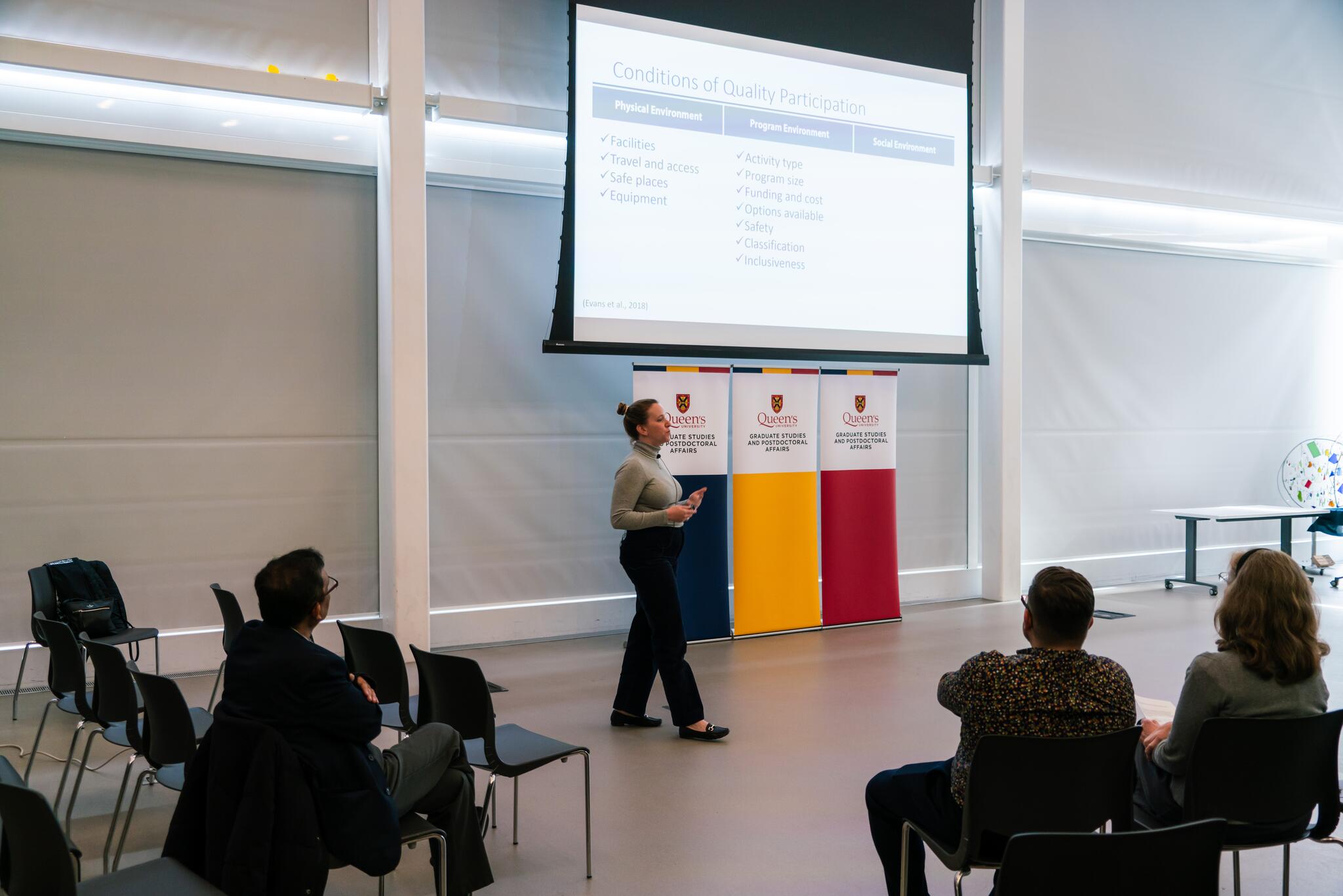 Woman in business casual standing in front of an audience that is sitting in chairs, taken from behind the audience. 