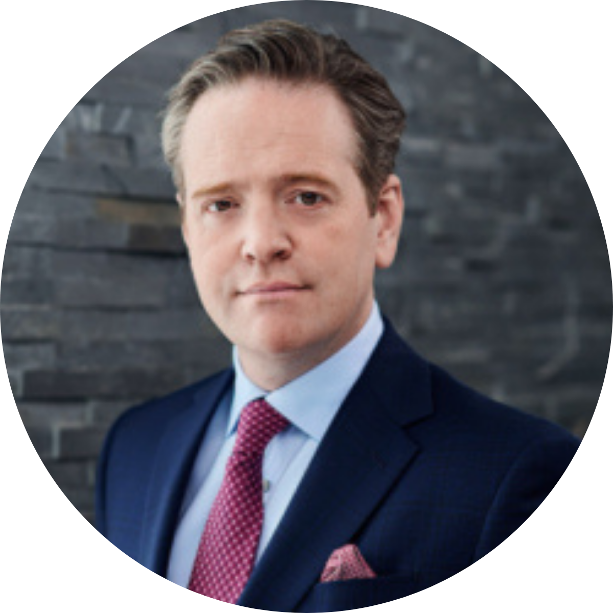 Man wearing navy blue suit and red tie with brown hair standing in front of a grey brick wall.